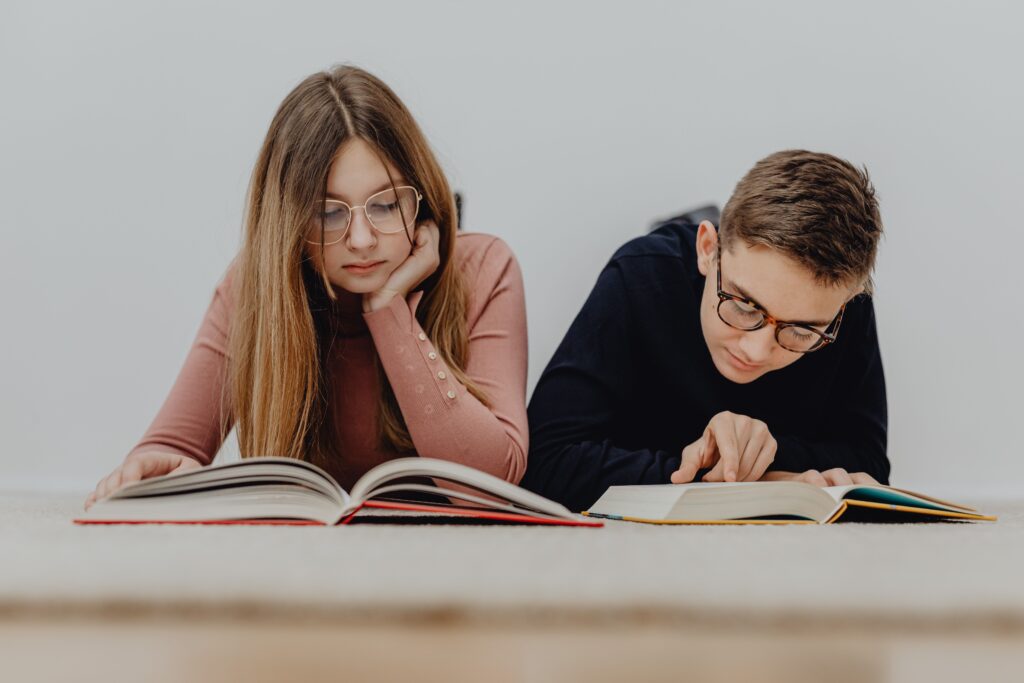 Two teenagers in eyeglasses reading books together indoors, focusing on learning and education.